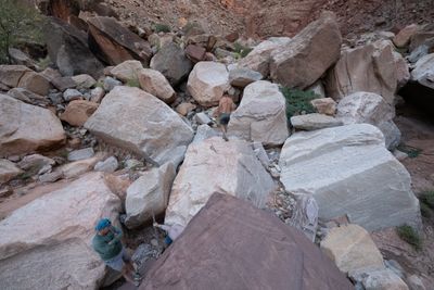 Seen from above, three men pick through a jumble of large pale boulders in a side canyon, one shirtless climbing ahead and one in a blue cap watching.