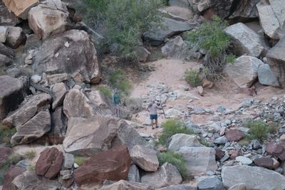 Telephoto view down onto two men standing in a sandy wash among large boulders and green shrubs, one holding a walking stick.