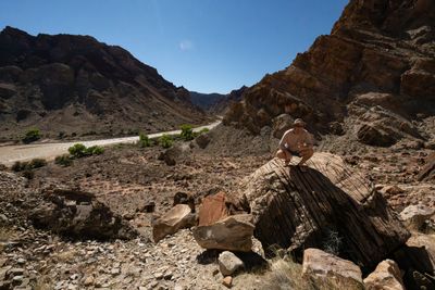 A man in a sun hat and sunglasses crouches on a large striated boulder on a rocky slope, the river winding past green riparian trees between dark canyon walls behind him.