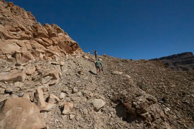 Two hikers climb a steep slope of loose scree below sandstone ledges, the lead one pointing upslope against a deep blue sky.