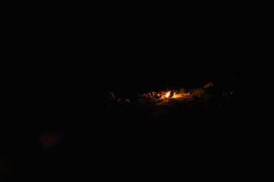 A small campfire glows among rocks in near-total darkness, a few faint points of light off to the right.