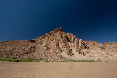 A jagged pinnacle of steeply tilted, fractured rock rises above a talus slope and a green band of riverside brush, flat brown water in front under a deep blue sky.