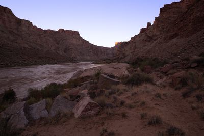 Wide view across a scrubby bench of boulders and dry grass down to a sand beach with camp gear and a white raft at the river's edge, the canyon rim catching first light.