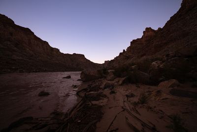 Before sunrise, silty river water laps a sand beach strewn with driftwood and boulders, between dark canyon walls under a pale lavender sky.