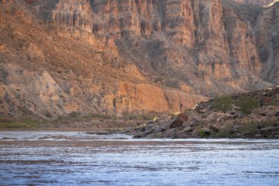 Nick, a small figure, stands on a large boulder on a rocky shoreline above riffled brown water, below tall sunlit canyon cliffs in warm morning light.
