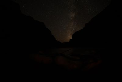 The Milky Way rises through a star-filled sky between black canyon walls, a small rock pinnacle silhouetted in the notch, rafts barely visible in the dark foreground.
