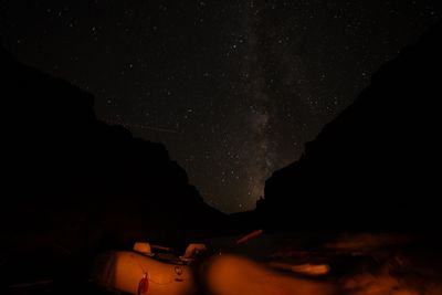 The Milky Way arcs between dark canyon walls over a small pinnacle in the notch, while warm light picks out a moored raft's tube, seats and oars in the foreground.