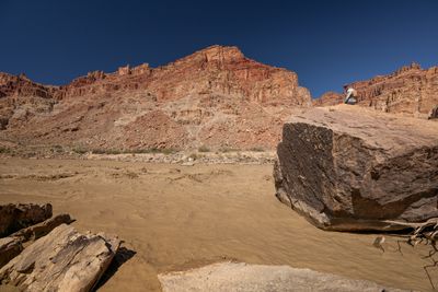 A person sits atop a huge angular boulder at the edge of a churning brown rapid, red layered cliffs and a deep blue sky behind.