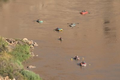 From high above, six loaded oar rafts and one small single-paddler boat drift spread out across calm brown water beside a grassy, rocky bank.