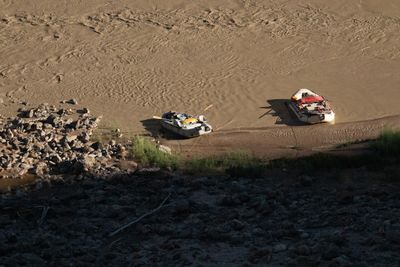 Looking straight down from a height, two loaded white rafts sit tied to a muddy shore in low golden light, casting long shadows on rippled brown water.