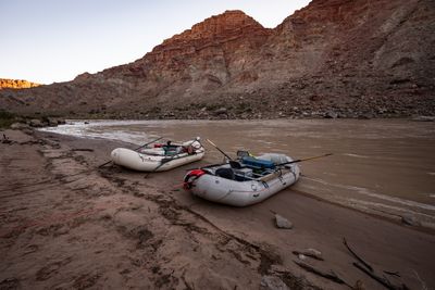 Two loaded, oar-rigged white rafts sit pulled up on a muddy sand beach, an orange bow line run up the shore, beside brown river water under a red layered cliff.