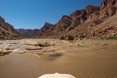 Over the white bow tube of a raft, brown water breaks into a riffle of small waves heading toward steep tilted cliffs and a boulder on the right shore.