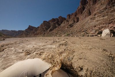Brown water splashes up over the white bow tube of a raft as it pushes through a wave, steep layered cliffs along the right bank under a deep blue sky.