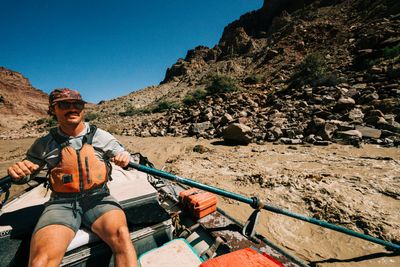 A man in a cap, sunglasses and orange life jacket pulls hard on the oars of a gear raft through churning brown waves beside a steep boulder slope.