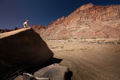 A person in a cap and life jacket sits on the top edge of a massive boulder in shadow, looking over a churning brown rapid toward sunlit red cliffs.