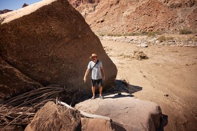 A man in a straw hat, T-shirt and shorts stands on a rock beside a giant boulder, pointing down and calling out, with a churning brown rapid behind him.