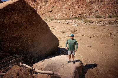 A smiling man in a blue cap, sunglasses and green long-sleeve shirt stands on a rock beside a giant boulder, can in hand, a churning brown rapid behind.