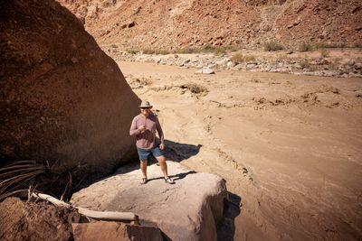 A man in a sun hat, sunglasses and mauve long-sleeve shirt throws a shaka on a rock beside a giant boulder, a churning brown rapid behind him.