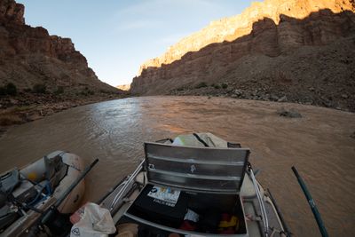A fisheye view from a raft's rowing seat over an open aluminum dry box and oar handles to a wide muddy river, with a second raft tied alongside and morning sun on the right canyon wall.