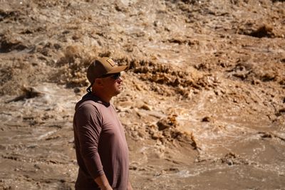 Profile of a man in a tan trucker cap with an orange patch, sunglasses and a maroon hooded sun shirt, standing in front of a wall of churning muddy whitewater.