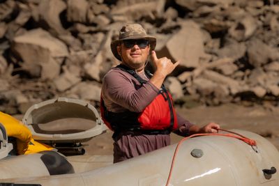 A wet, grinning man in a bucket hat, sunglasses and a red life jacket flashes a shaka from a white raft, holding the bow line, with sunlit boulders blurred behind.