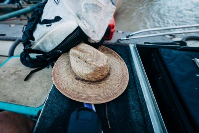 A sweat-stained palm-leaf sun hat rests on the black carpeted deck of a raft beside a white backpack, an open aluminum dry box and a lens cap, with muddy river water at the corner.