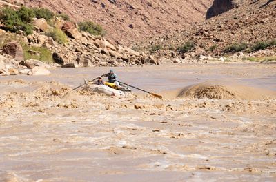 An oarsman rows a loaded white gear raft into a muddy rapid, a passenger seated up front, as a large brown wave rears to the right below a boulder-covered bank.
