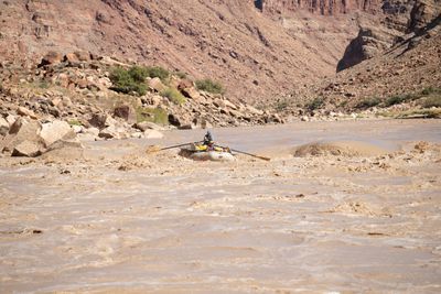 A loaded white gear raft with an oarsman and a passenger works through a churning muddy rapid beside a big brown wave, below boulder banks and red canyon walls.