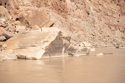 A huge cracked sandstone block with a dark water-stained base sits at the edge of a calm muddy river below a talus slope and cliff, with a small raft and its rower far off to the right.