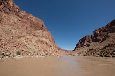 A single raft floats far downstream on a broad, calm muddy river between towering red layered canyon walls under a deep blue cloudless sky.