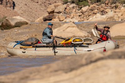 Side view of a loaded white gear raft among boulders: an oarsman in a plaid shirt and teal cap sits at the oars as a passenger in a red life jacket holds up a white bag and flashes a shaka.