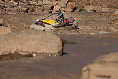 An oarsman in a teal cap and life jacket rows a loaded white raft through brown whitewater as a passenger in a bucket hat grips the bow line, past an out-of-focus boulder in front.