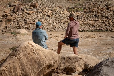 Two men seen from behind on pale sandstone boulders watch a churning muddy rapid: one in a teal cap and plaid shirt with arms folded, one in a bucket hat and maroon hoodie with a foot propped up.
