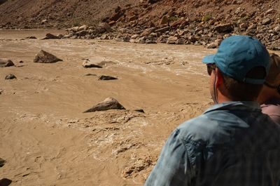Over the shoulder of a man in a teal cap and blue shirt, with a second person beside him, looking down on a muddy rapid scattered with rocks below a boulder bank.