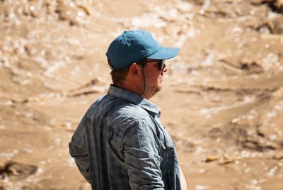 Profile of a man in a teal ball cap, sunglasses on a retainer cord and a gray plaid shirt, looking upstream at out-of-focus churning muddy whitewater.