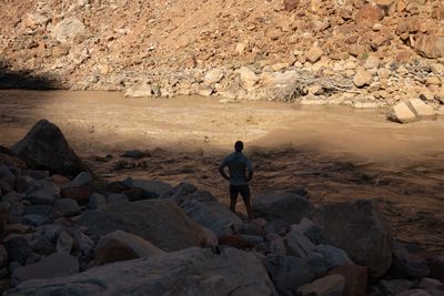 A man in silhouette stands with his hands on his hips on shaded boulders, looking at a muddy rapid, with a sunlit talus slope and a driftwood pile across the river.
