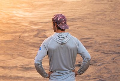 Seen from behind, a man in a backward floral cap and a pale gray hooded sun shirt stands with his hands on his hips, facing rippled muddy river water glowing gold in the sun.