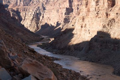 From a talus slope high above, a muddy river winds through a deep canyon of stepped red sandstone cliffs, half in shadow and half lit by low morning sun.