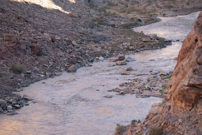 An elevated view down onto a muddy river running through a boulder-strewn rapid, with two rafts tied at a small sandy camp, gear spread on the beach, and a sunlit rock face in the foreground.