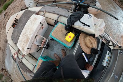 Fisheye view straight down at bare, muddy feet on a raft's diamond-plate floor among a white cooler, teal seat boxes, a yellow hard case, a straw hat and a backpack.
