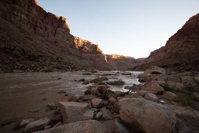 A low wide-angle view from a boulder shore across a muddy river riffling through rocks, with a driftwood snag in front and early sun lighting the tops of tall red canyon walls under a clear sky.
