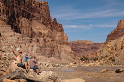 Three smiling men sit on a sandstone boulder beside a muddy rapid, waving and holding cans, below towering red layered cliffs, a rock pinnacle and blue sky.