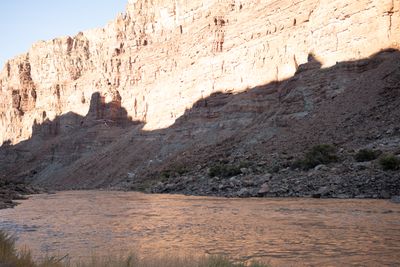 Two small ultralight trike aircraft fly low past a towering sunlit sandstone wall, above a talus slope in shadow and a wide muddy river in the foreground.