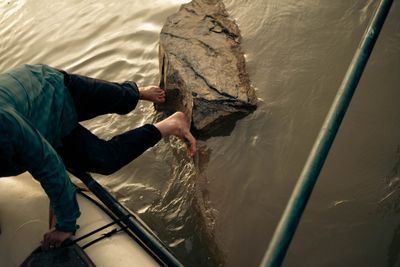 Looking down from a raft as a barefoot boatman braces both feet against a mid-river boulder to push the boat off in shallow, silty water.