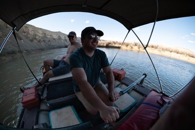 An oarsman rows a loaded raft beneath a bimini shade canopy on calm water, canyon walls behind.