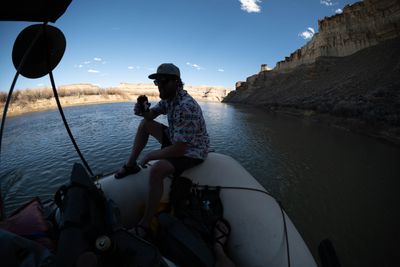 A crew member sits silhouetted on the bow tube against canyon walls and a cloud-streaked afternoon sky.