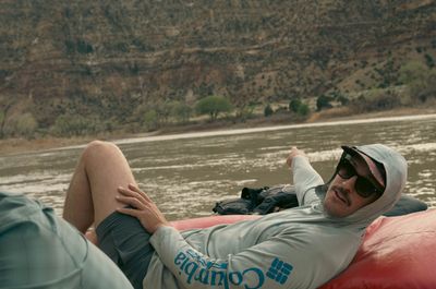 Crew member stretched out on the bow tube of a loaded raft drifting calm brown water.