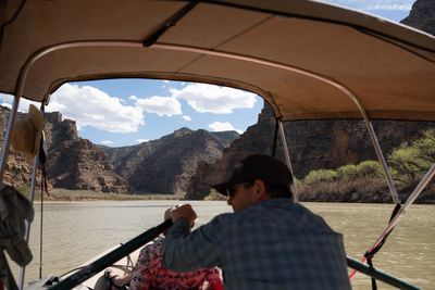 Two crew members under the raft's shade canopy, rowing toward layered canyon walls.