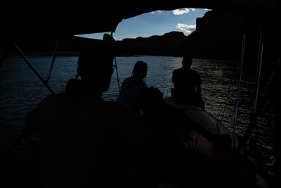 Three rafters sit in near-total silhouette under a bimini shade canopy on a calm stretch of the Green River at dusk, with dark canyon walls and a narrow strip of pale blue sky framing the water ahead.