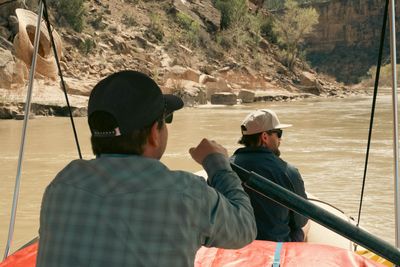 Two rafters under the boat frame, one at the oars, drifting silty water past a boulder bank below layered canyon walls.